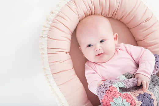 Baby som ligger i babynest med heklet teppe over seg og smiler